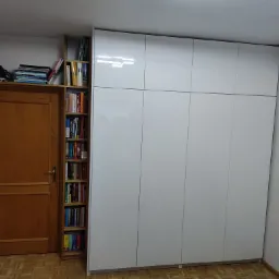 Biała szafa wnękowa z połyskiem, przylegająca do drewnianego regału z książkami i drewnianych drzwi z książkami na górze, na parkiecie.