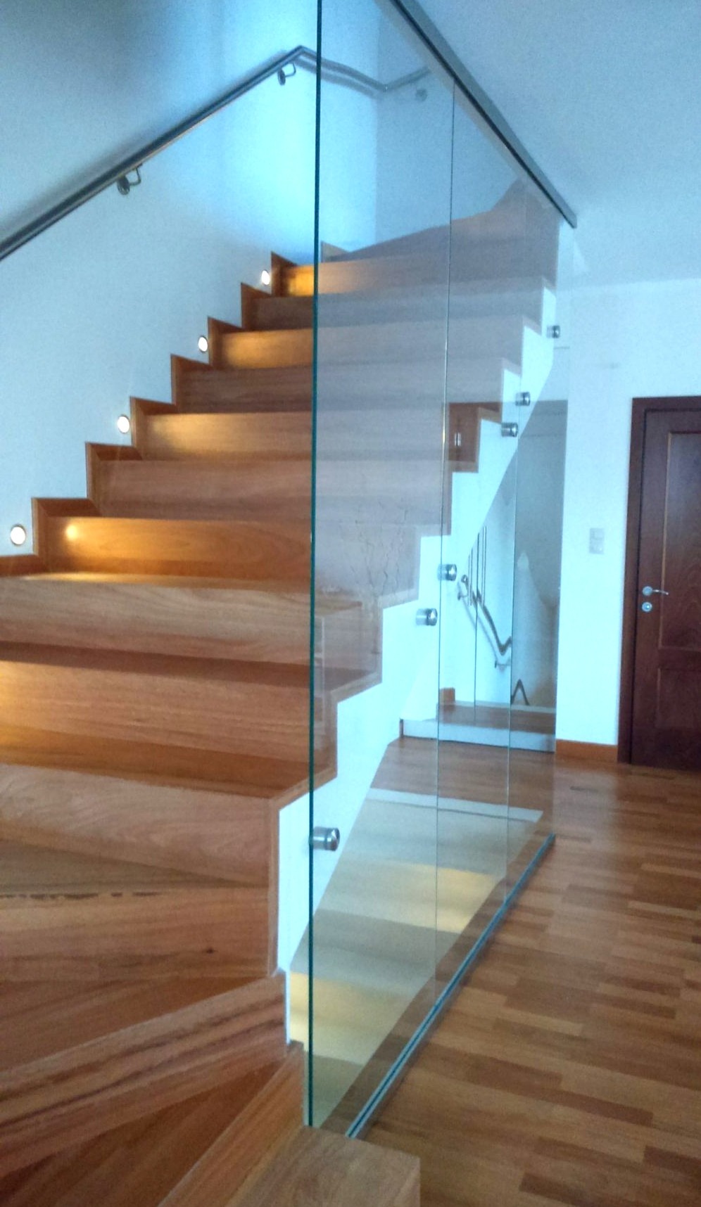 Drewniane schody z oświetleniem punktowym na stopniach i szklaną balustradą, widoczne drewniane podłogi i drzwi.