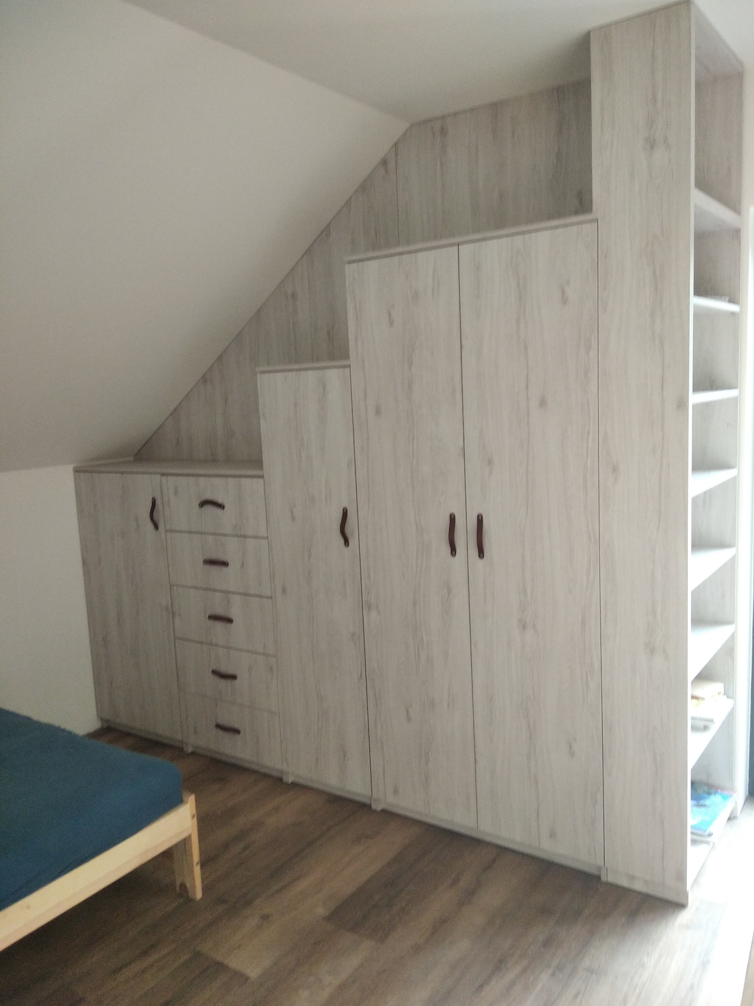 Zabudowa wnęki na poddaszu z szafami i szufladami, wykończona laminatem imitującym drewno, z widocznym fragmentem łóżka.