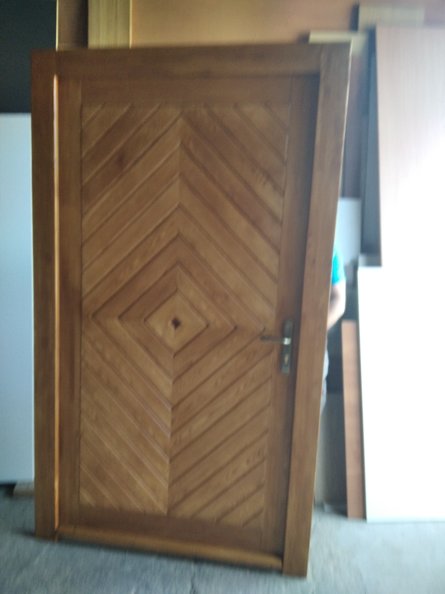 Drewniane drzwi wewnętrzne z geometrycznym wzorem w romby, widoczne z bliska, z klamką i fragmentem osoby w tle.