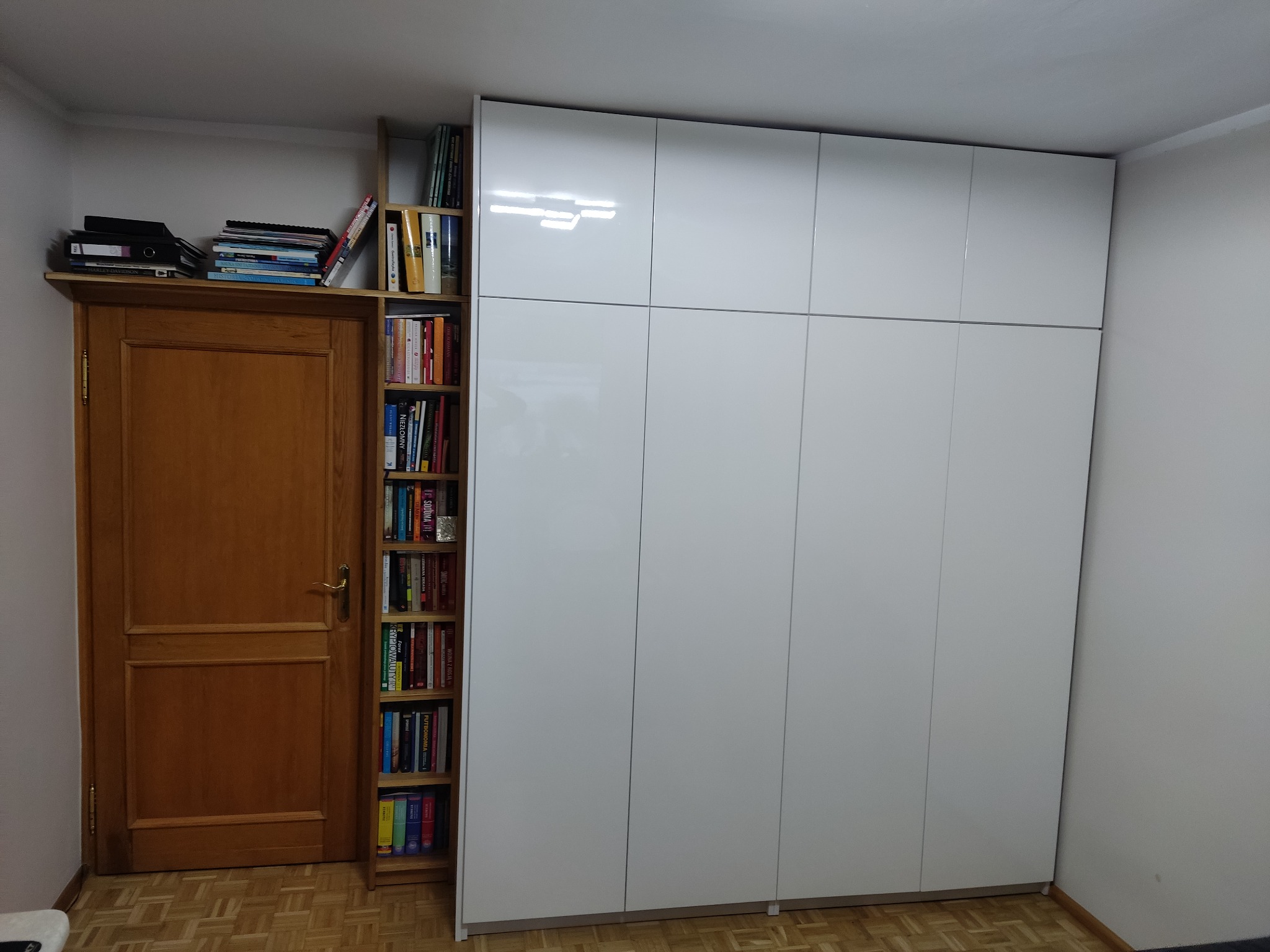 Biała szafa wnękowa z połyskiem, przylegająca do drewnianego regału z książkami i drewnianych drzwi z książkami na górze, na parkiecie.