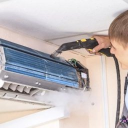 Trodos Instalacje - Mężczyzna w kombinezonie roboczym czyści parą wnętrze klimatyzatora ściennego, widoczne niebieskie lamele i wydobywająca się para.