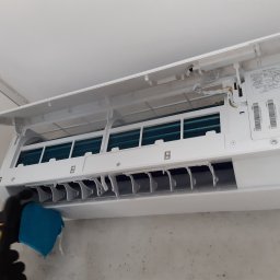 Trodos Instalacje - Czyszczenie wewnętrznych elementów klimatyzatora ściennego przez osobę w rękawicach roboczych, widoczne niebieskie lamele chłodzące i zbliżenie na czyszczoną kratkę wylotową powietrza.