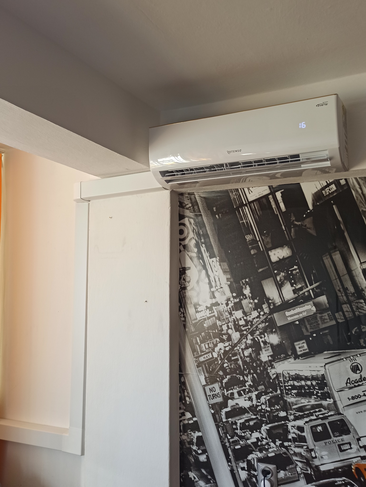 Biały klimatyzator marki Rotenso zamontowany w pomieszczeniu z fototapetą przedstawiającą ruchliwą ulicę Nowego Jorku, widoczny wyświetlacz z temperaturą 16 stopni.