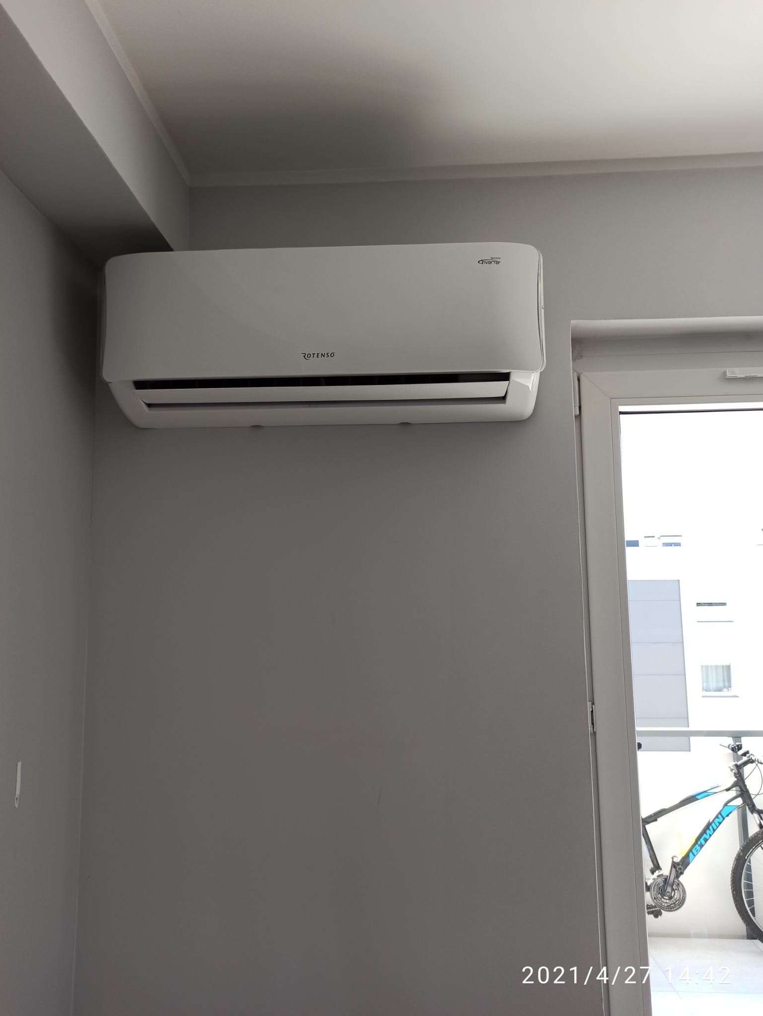 Biały klimatyzator marki Rotenso zamontowany na szarej ścianie wewnątrz pomieszczenia, z widocznym fragmentem roweru na balkonie w tle.