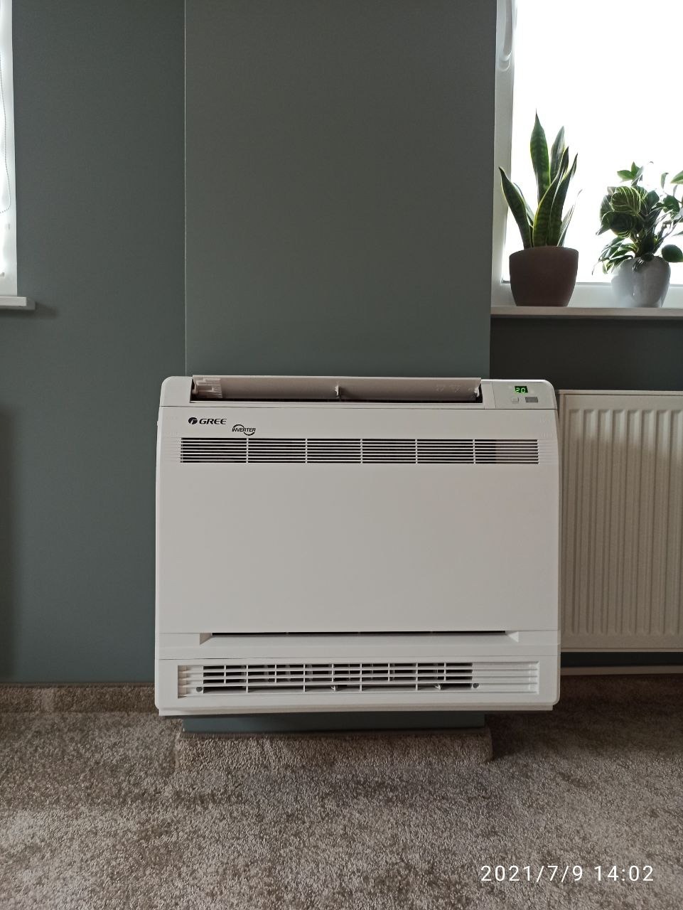 Biały klimatyzator podłogowy marki Gree Inverter w nowoczesnym wnętrzu z szarą ścianą, obok okna z roślinami doniczkowymi.