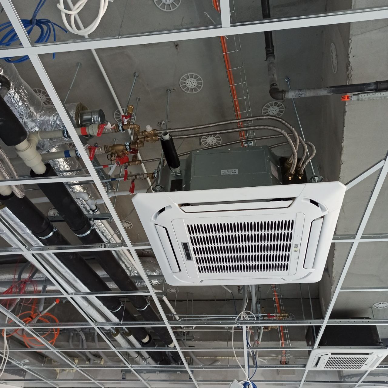 Kasetonowa jednostka klimatyzacyjna zamontowana pod sufitem rastrowym, widoczne rury instalacyjne i okablowanie.