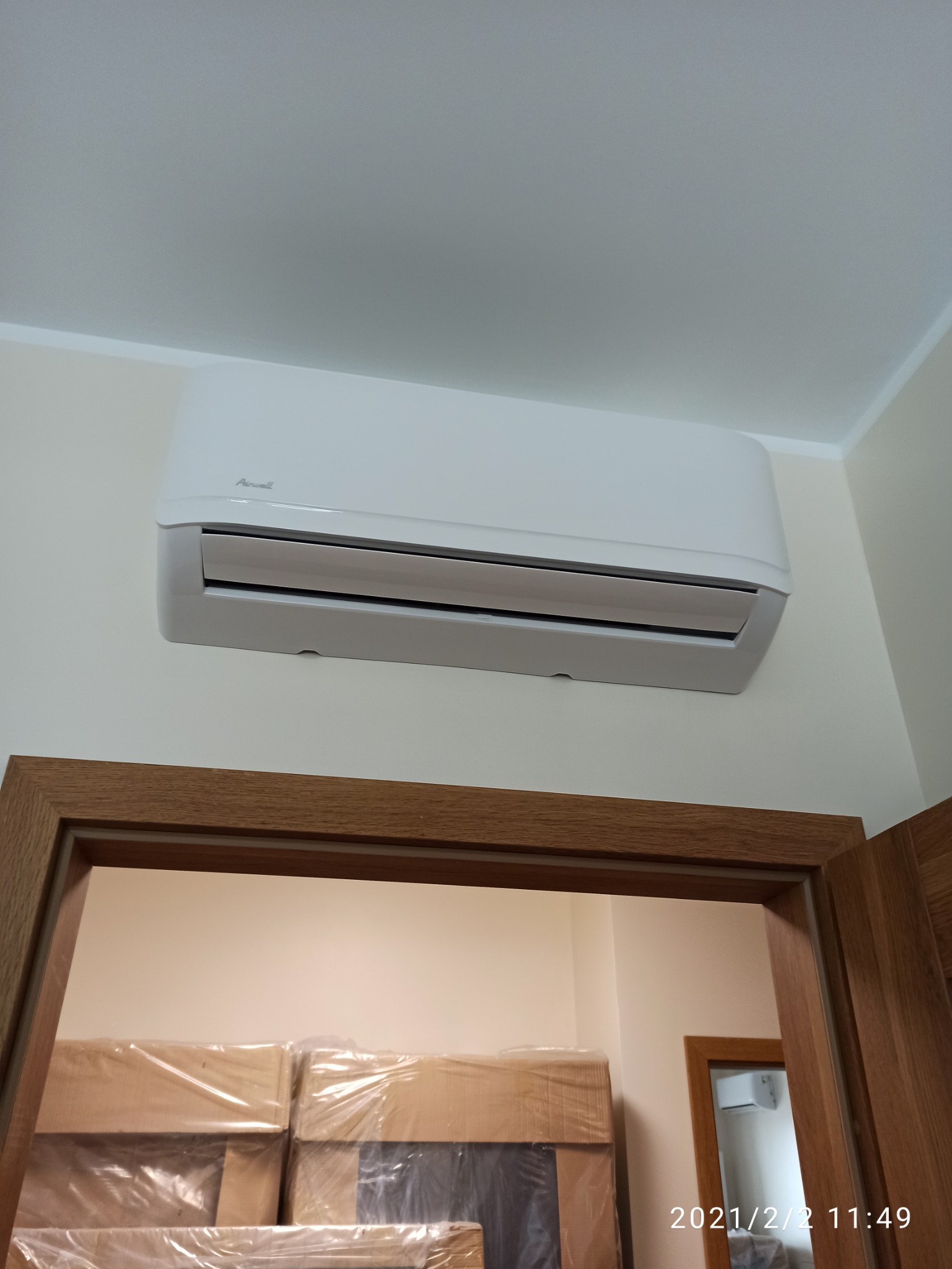 Biały klimatyzator marki Airwell zamontowany na ścianie nad drewnianą futryną drzwi. W tle widoczne zapakowane kartony.