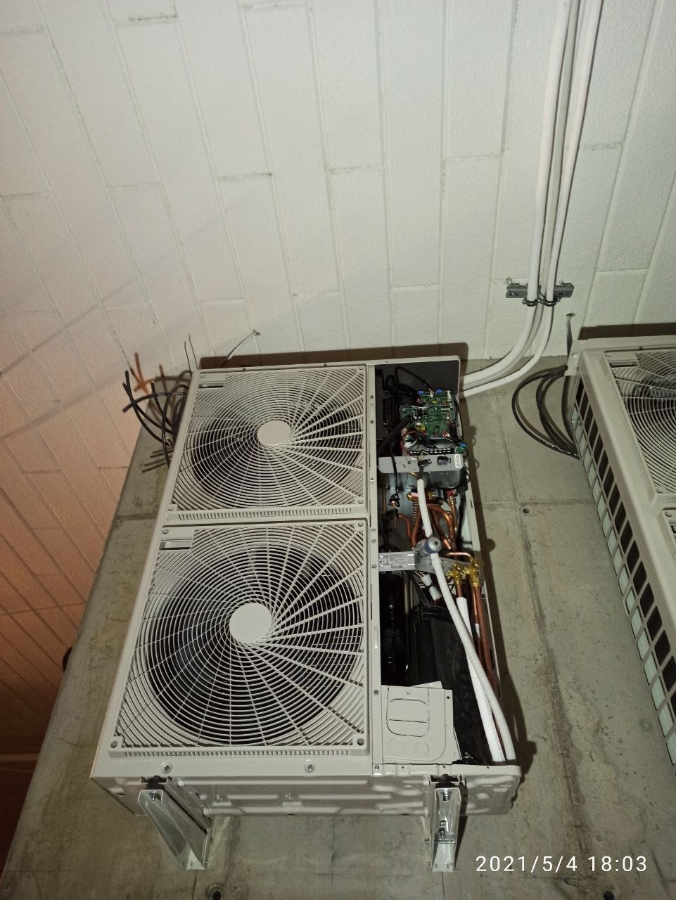 Zewnętrzna jednostka klimatyzacji z otwartą obudową, widoczne elementy wewnętrzne, instalacja elektryczna i rury chłodnicze, zamontowana na betonowej ścianie z widocznymi przewodami.