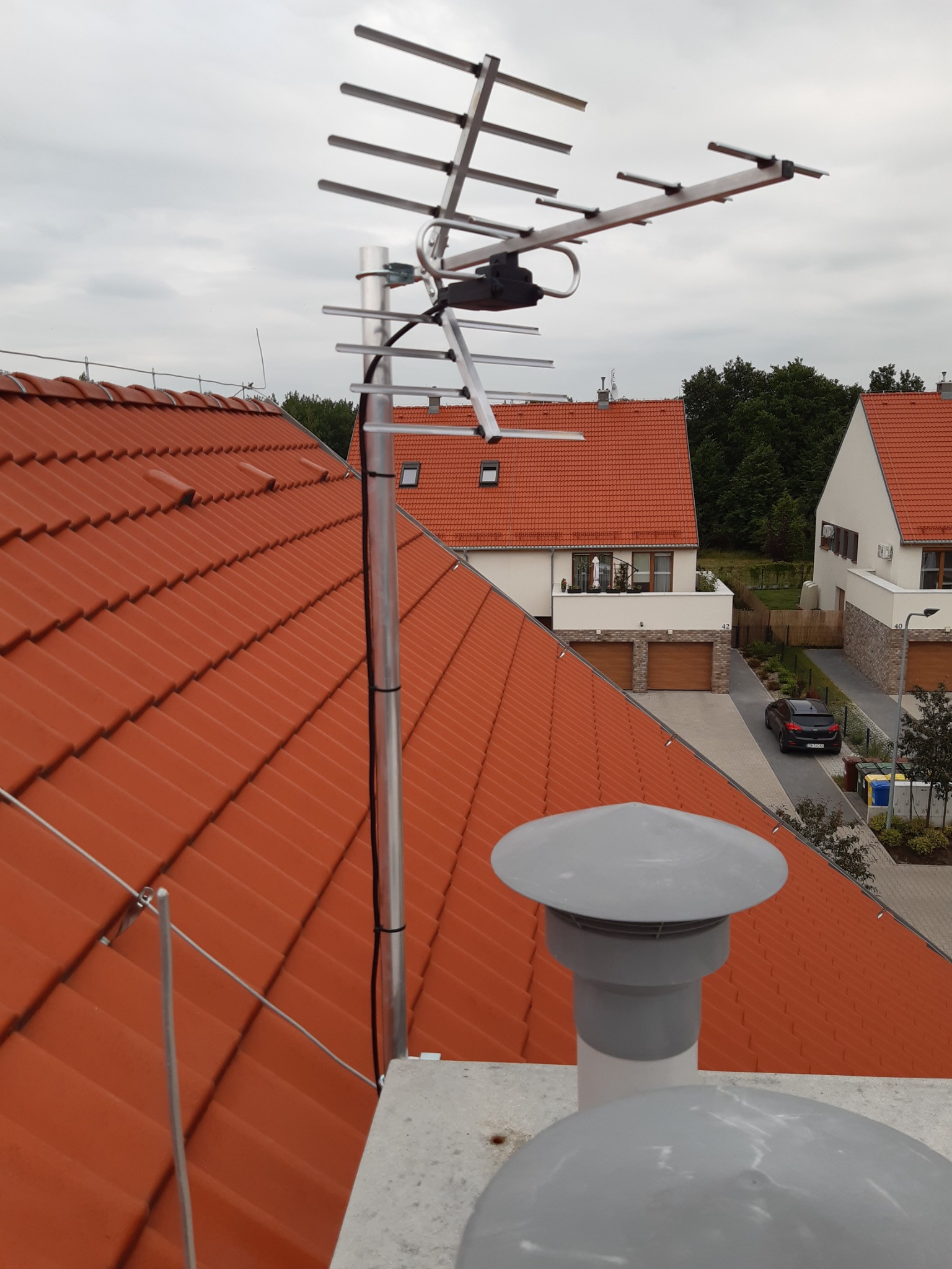 Antena telewizyjna zamontowana na dachu domu z czerwoną dachówką, w tle widoczne inne domy i pochmurne niebo.