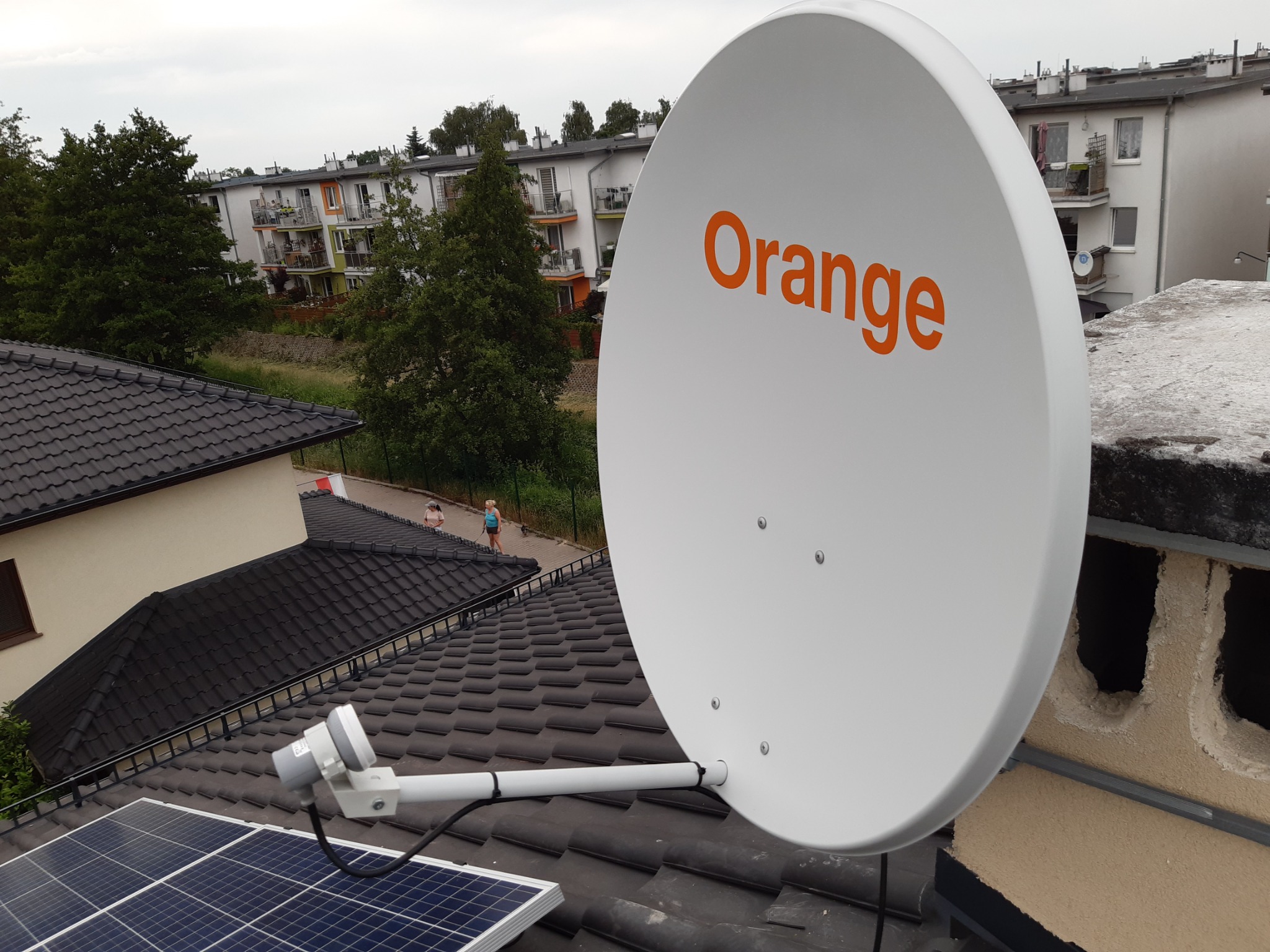 Biała antena satelitarna z pomarańczowym logo na dachu z panelami słonecznymi, widok na osiedle mieszkaniowe w tle.