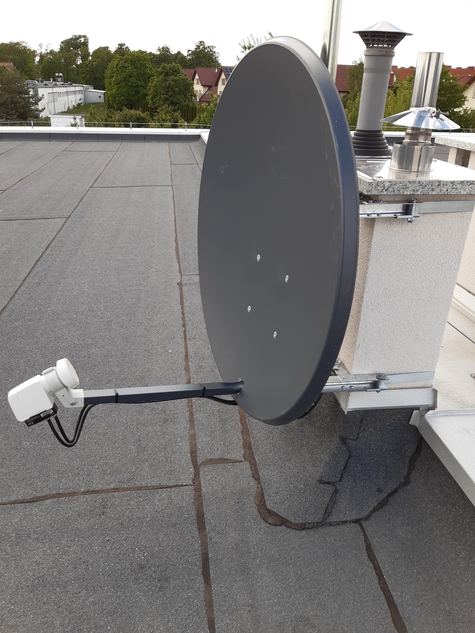 Szara antena satelitarna zamontowana na płaskim dachu z papy, przymocowana do komina za pomocą metalowych wsporników.