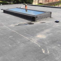 Fortis Marcin Wawrzeńczak - Płaski dach pokryty czarną membraną bitumiczną z świetlikiem dachowym i kominkami wentylacyjnymi, widoczne ślady po wodzie i zabrudzenia.