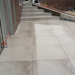 Fortis Marcin Wawrzeńczak - Nowoczesny taras z szarych, kwadratowych płyt, częściowo mokrych, prowadzący do schodów i budynku z dużymi oknami, widoczne prace wykończeniowe.