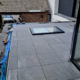 Szare płytki na dachu tarasu z oknem dachowym, widoczny fragment budynku z cegły i nowoczesna bryła domu, niebieska folia zabezpieczająca krawędź dachu.