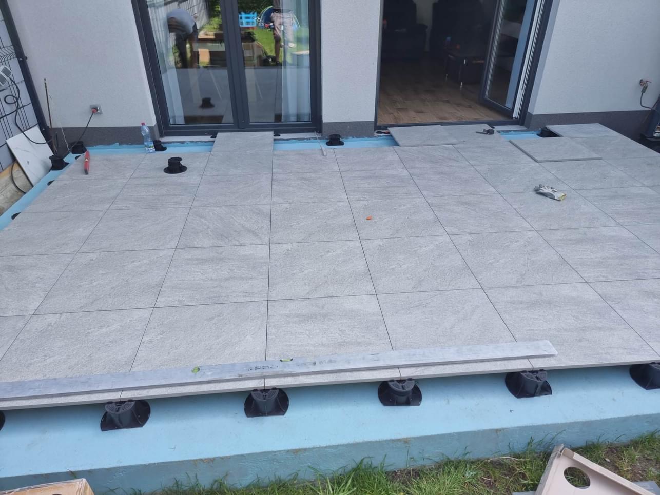 Układanie szarych płyt betonowych na regulowanych wspornikach tarasowych, widoczny poziomica i częściowo ułożona powierzchnia tarasu przed budynkiem.