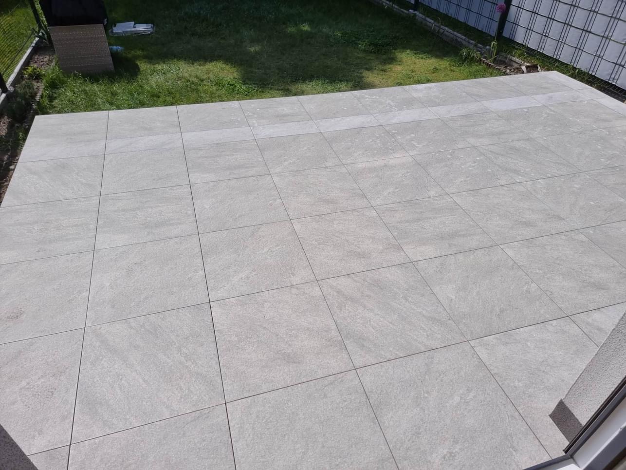 Szary taras z płytek imitujących kamień, widok z góry na trawnik i ogrodzenie panelowe.