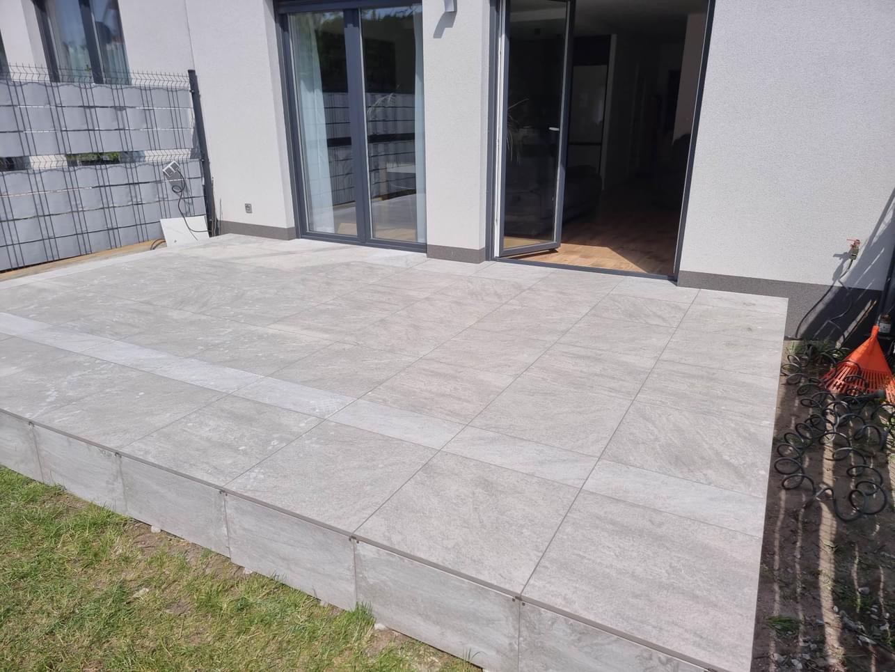 Nowoczesny taras wyłożony szarymi płytami, z widocznymi drzwiami przesuwnymi prowadzącymi do wnętrza domu i fragmentem trawnika z boku.