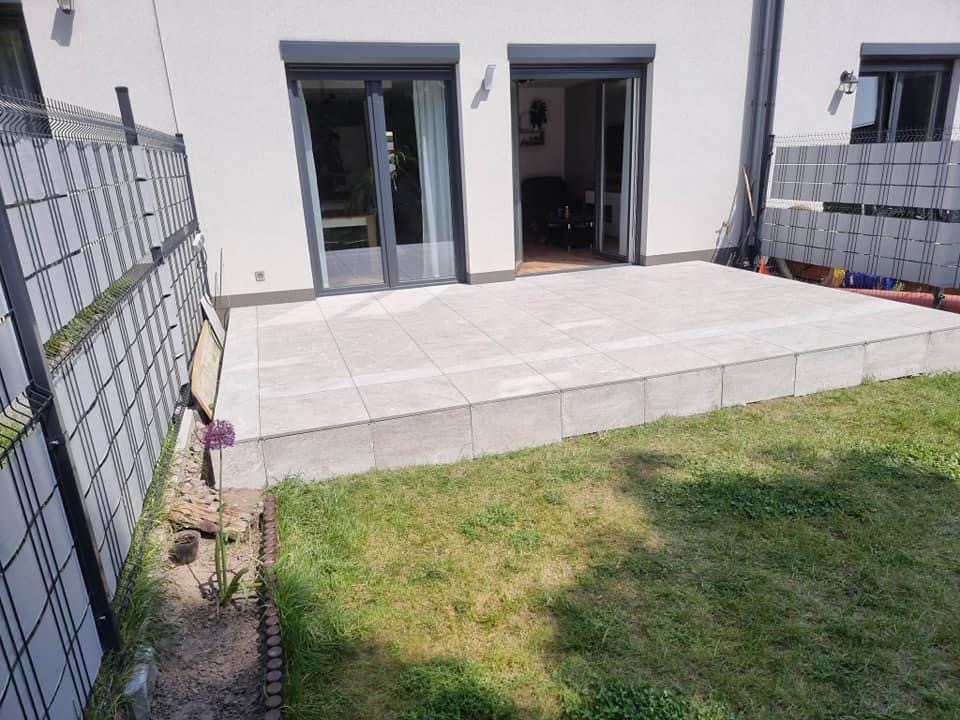 Nowoczesny taras wyłożony jasnymi płytami betonowymi, przylegający do białego budynku z przesuwnymi drzwiami tarasowymi, widoczny fragment trawnika i ogrodzenia panelowego.