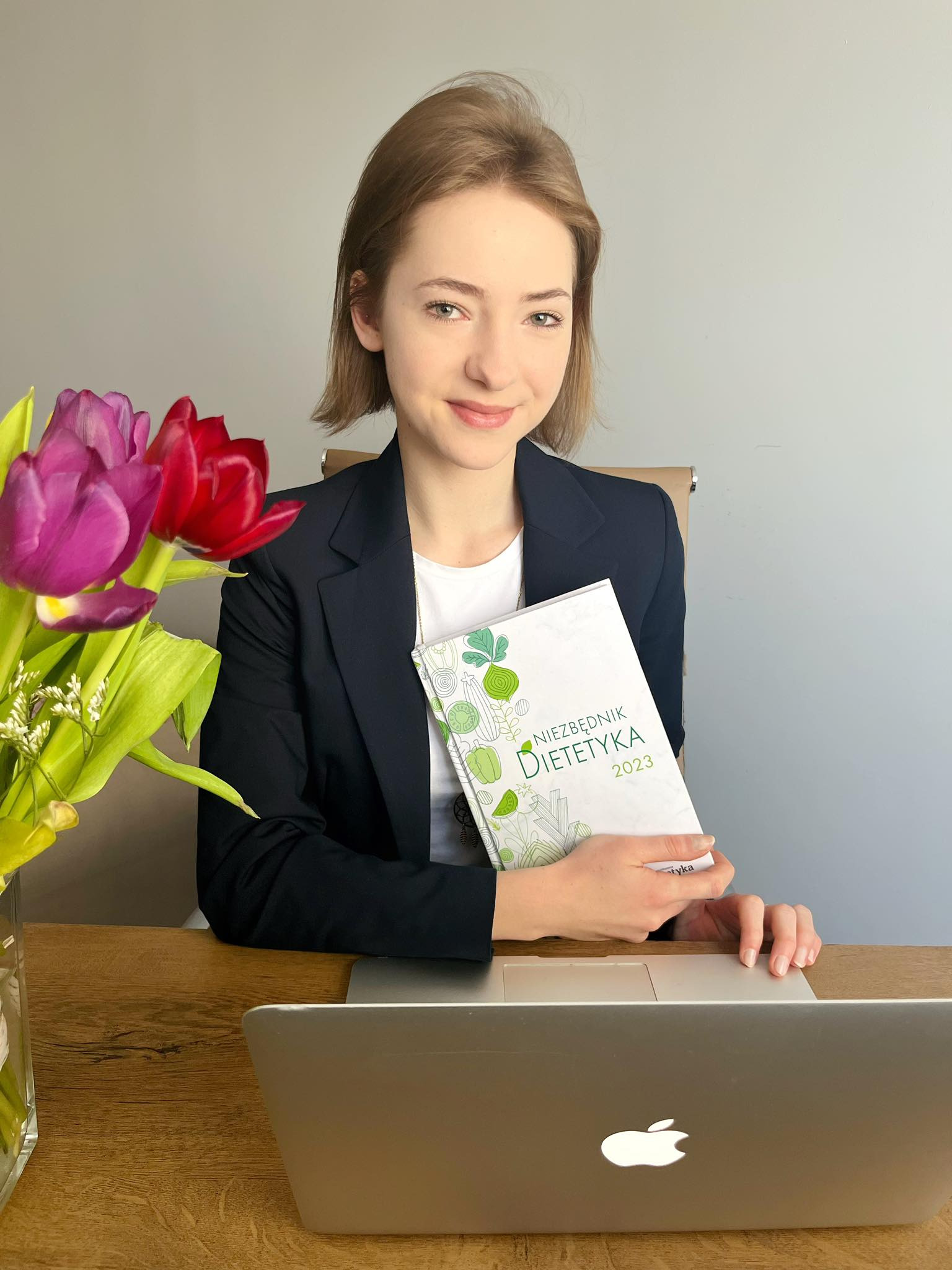Uśmiechnięta kobieta w marynarce trzymająca kalendarz 'Niezbędnik Dietetyka 2023' przy laptopie, obok w wazonie tulipany.