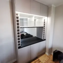 Jasnoszara szafa wnękowa z czarnym blatem i oświetleniem LED, z półkami na wino po bokach, odbijająca fragment sofy w lustrzanej wnęce, na tle drewnianej podłogi.