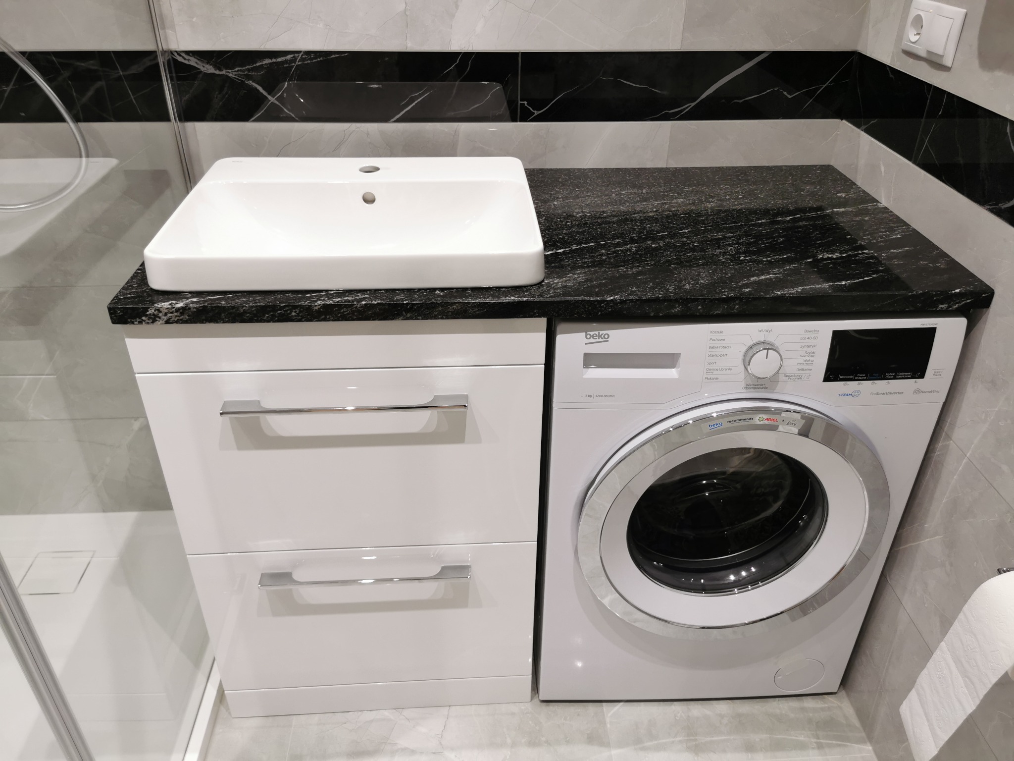 Biała umywalka nablatowa na blacie z ciemnego kamienia, po prawej pralka Beko, całość w łazience wykończonej szarymi płytkami z czarnym pasem dekoracyjnym.