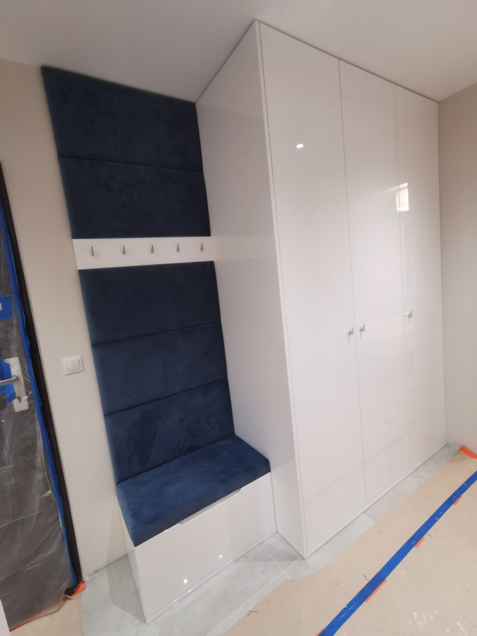 Biała szafa wnękowa z siedziskiem i panelem tapicerowanym w kolorze granatowym, na którym zamontowane są wieszaki; podłoga zabezpieczona kartonem i taśmą malarską.