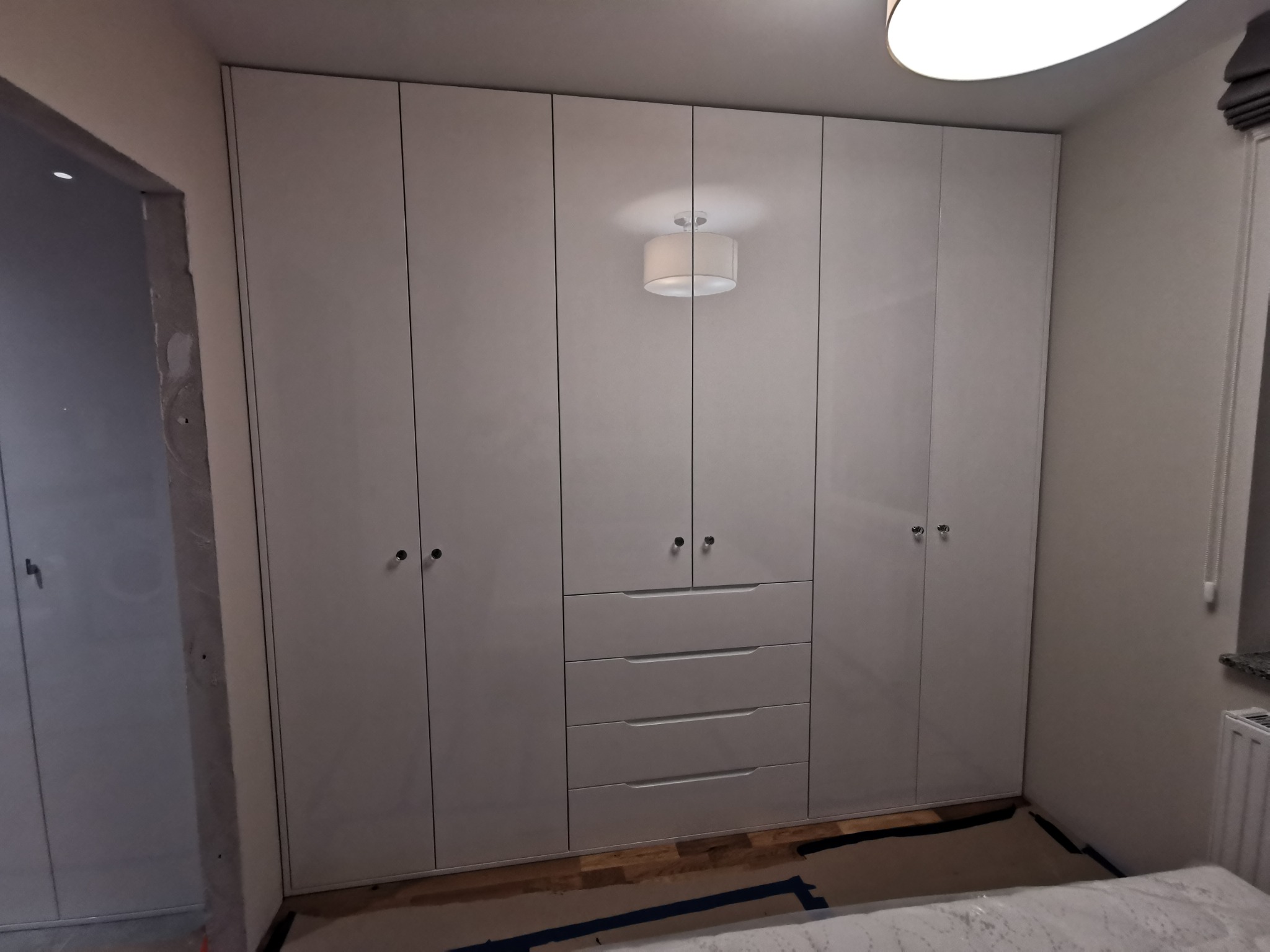 Jasnoszara szafa wnękowa z szufladami i okrągłymi uchwytami, zamontowana od podłogi do sufitu w mieszkaniu w Sycowie, widoczny fragment podłogi zabezpieczony kartonem.