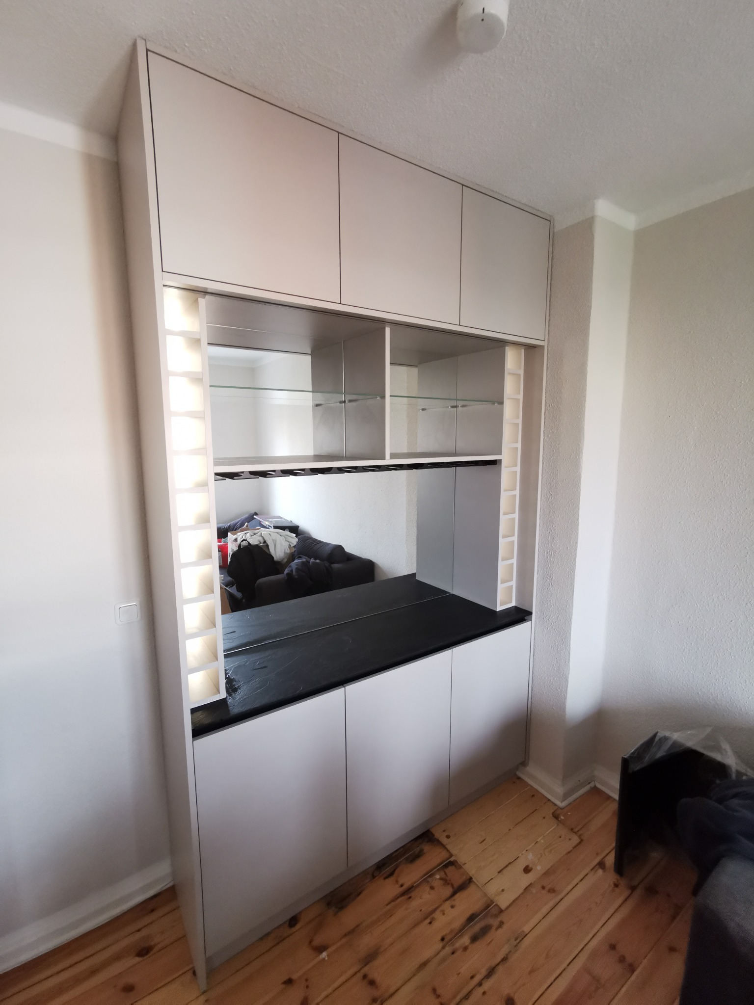 Biała zabudowa meblowa z czarnym blatem i lustrzanymi półkami, zintegrowana z oświetleniem LED, na tle drewnianej podłogi.