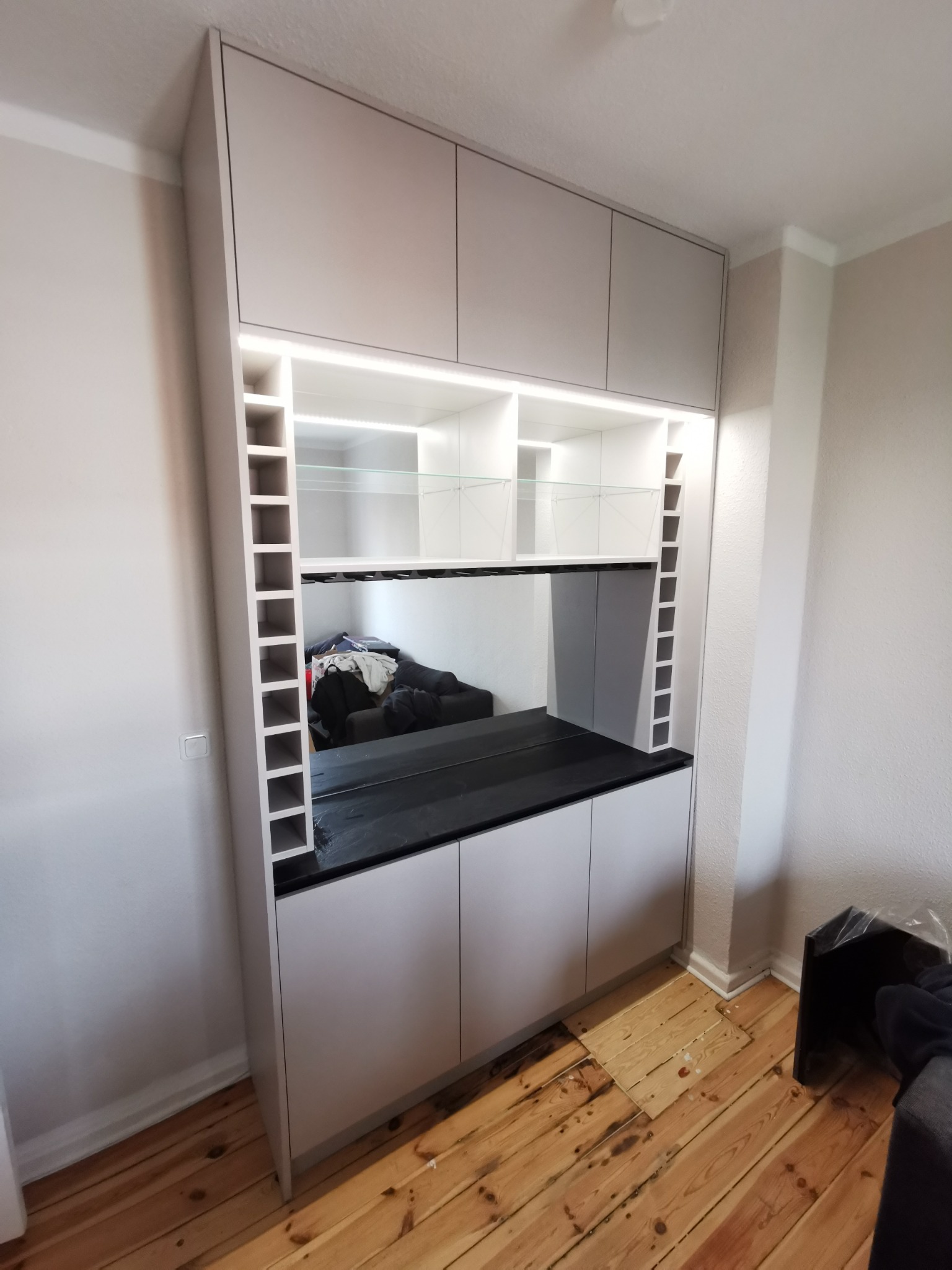 Jasnoszara szafa wnękowa z czarnym blatem i oświetleniem LED, z półkami na wino po bokach, odbijająca fragment sofy w lustrzanej wnęce, na tle drewnianej podłogi.