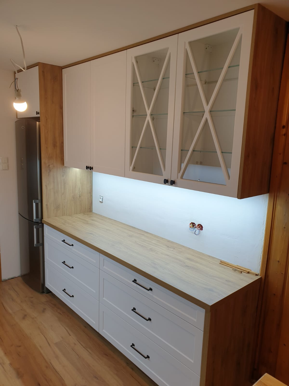 Nowoczesna kuchnia z białymi szafkami dolnymi i górnymi z przeszkleniami w kształcie krzyży, drewniany blat, podświetlenie LED oraz widoczne gniazdko elektryczne w trakcie montażu.