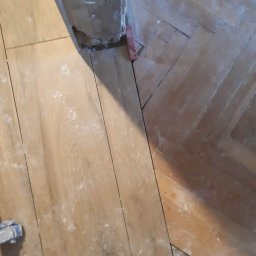 Babik Wykończenia Wnętrz - Fragment podłogi z widocznym połączeniem nowych paneli i starego parkietu w jodełkę, z widocznymi śladami pyłu i czerwonymi klinami dystansowymi.