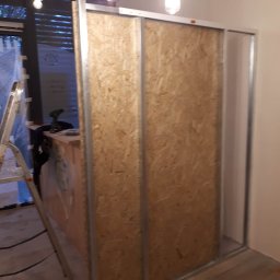 Babik Wykończenia Wnętrz - Wnętrze w trakcie remontu: konstrukcja ścianki działowej z płyty OSB na metalowym stelażu, widoczna drabina i narzędzia w tle, surowe oświetlenie.