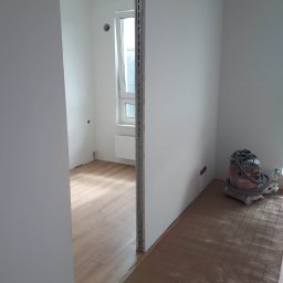 Babik Wykończenia Wnętrz - Wnętrze w trakcie remontu: świeżo pomalowane białe ściany, widoczny fragment podłogi z ogrzewaniem podłogowym i odkurzacz przemysłowy w rogu pomieszczenia.