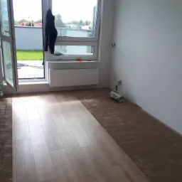 Wnętrze pokoju w trakcie układania paneli podłogowych, widoczna część ułożonej podłogi z jasnych paneli oraz fragment przygotowanego podłoża z brązowej maty, otwarte drzwi balkonowe z widokiem...