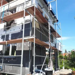 Dom w trakcie ocieplania, widoczne rusztowania, czarny styropian i szara warstwa kleju, elementy ocieplenia leżą na paletach przed budynkiem.