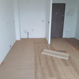 Układanie paneli podłogowych w jasnym wnętrzu, widoczne przygotowane podłoże, fragment listwy przypodłogowej i instalacja wodna na ścianie.