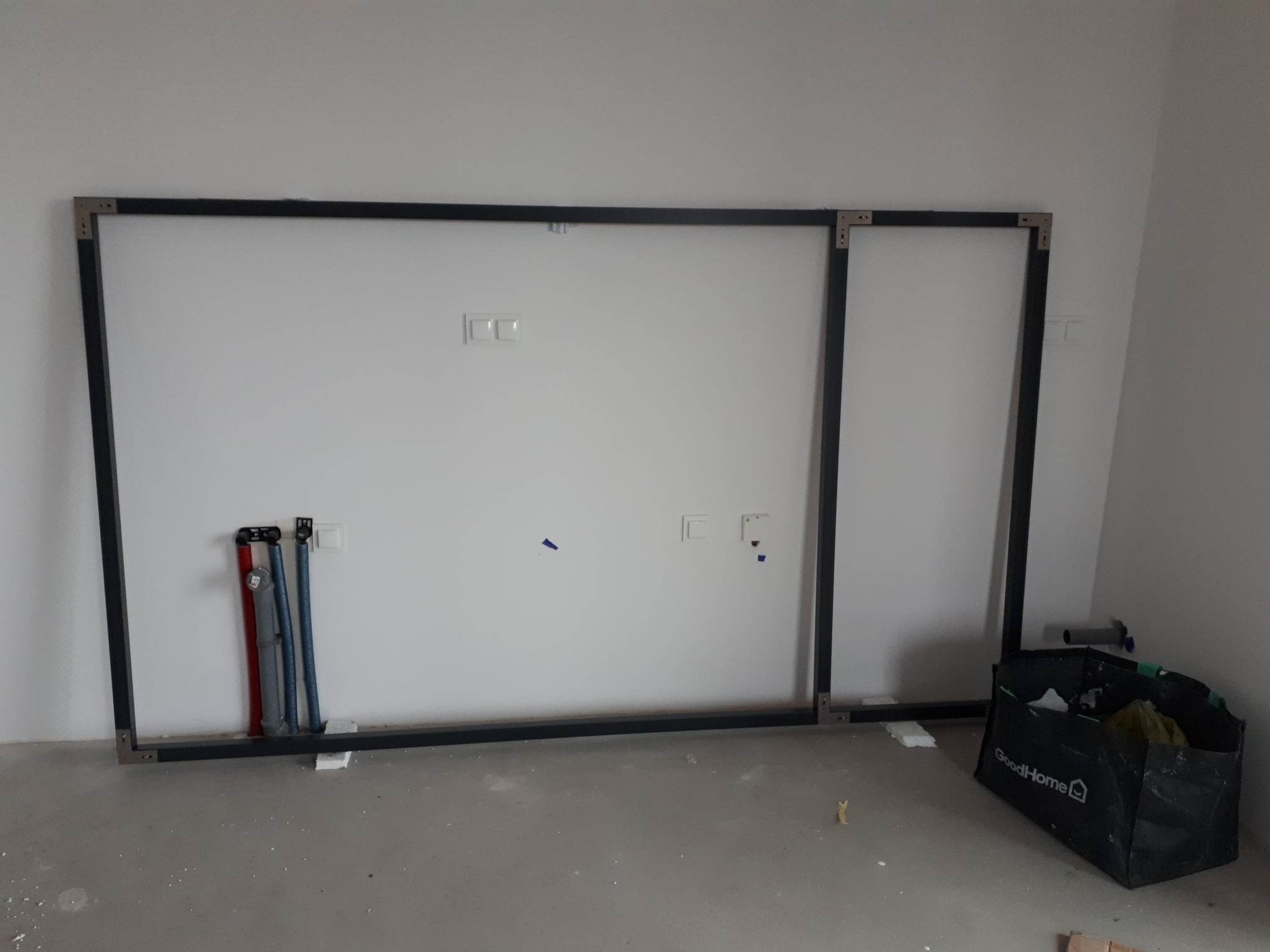 Stelaż z profili metalowych przy białej ścianie z instalacją elektryczną i hydrauliczną, torba budowlana GoodHome na podłodze.