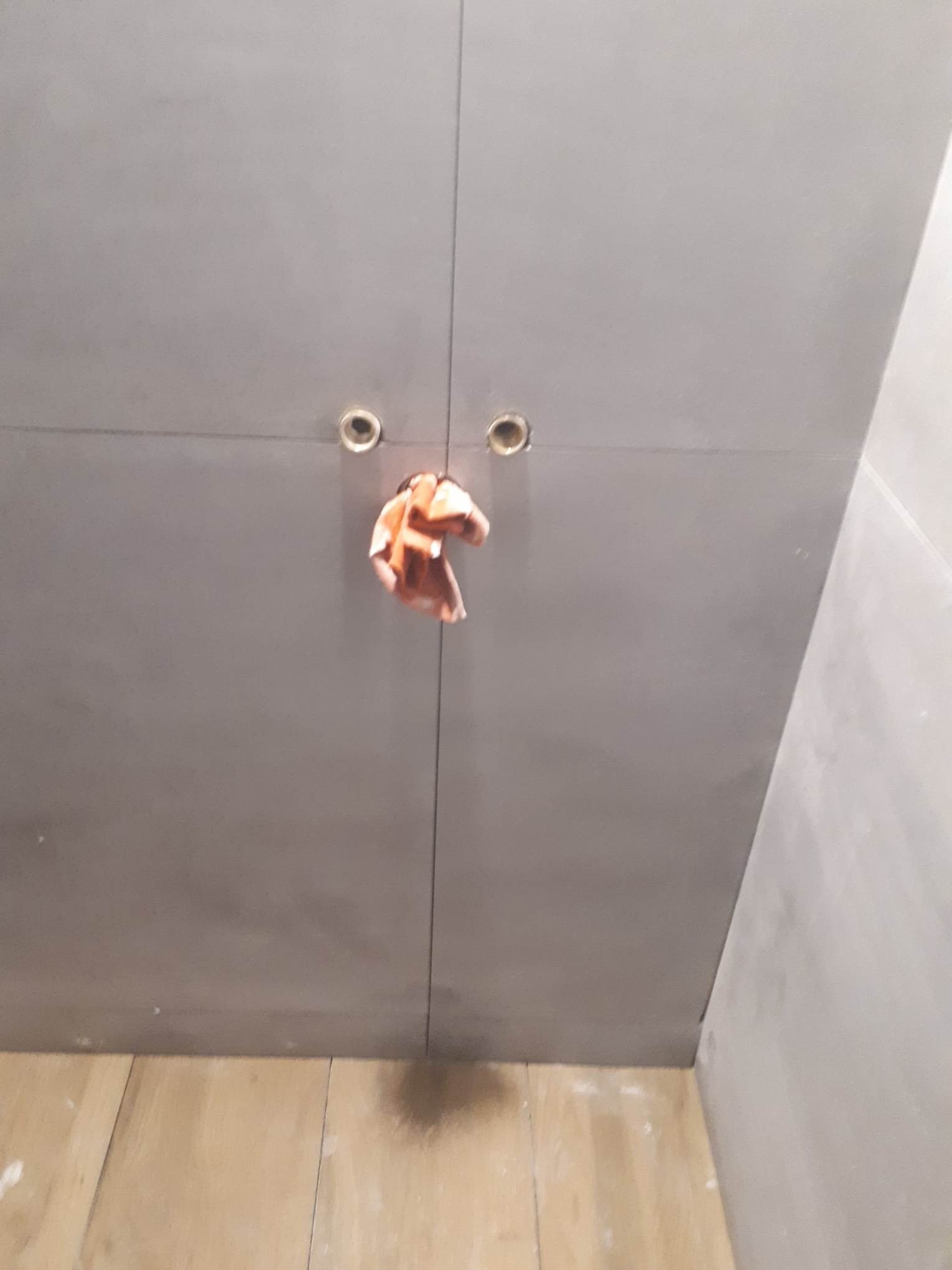 Szare, matowe drzwi szafy z dwoma otworami na uchwyty, z których wystaje pomarańczowa szmatka, drewniana podłoga z widocznymi śladami po remoncie.