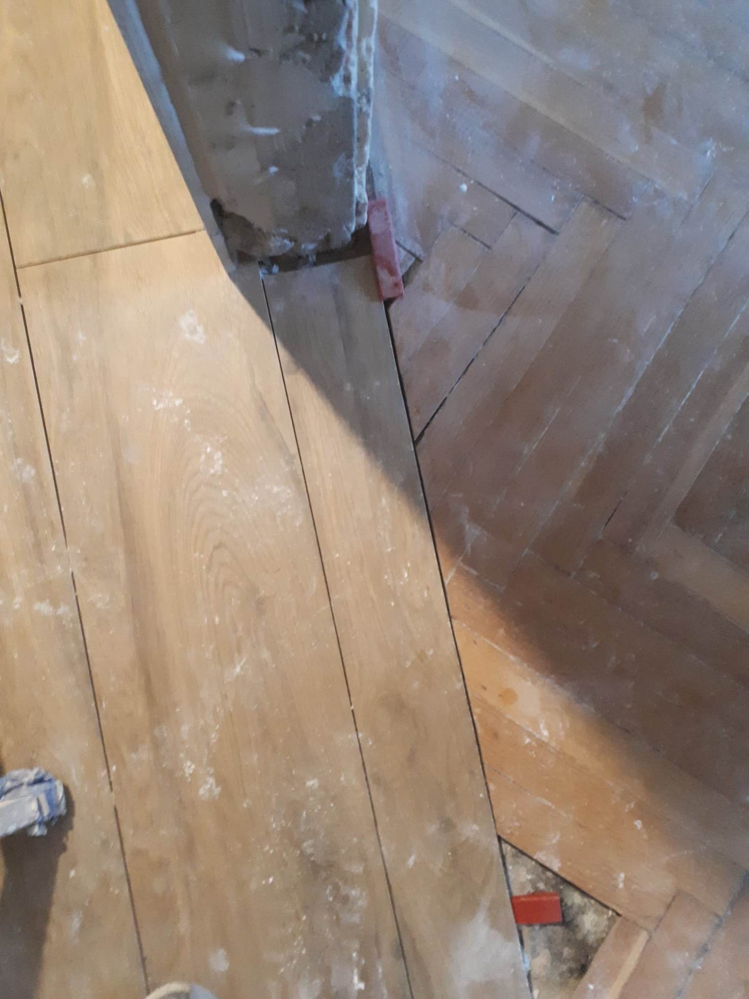 Fragment podłogi z widocznym połączeniem nowych paneli i starego parkietu w jodełkę, z widocznymi śladami pyłu i czerwonymi klinami dystansowymi.