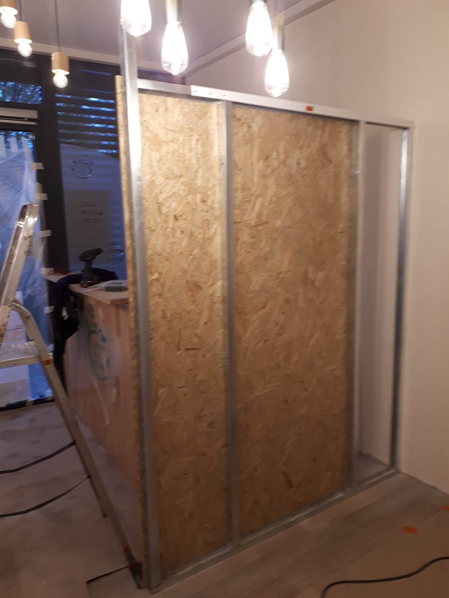 Wnętrze w trakcie remontu: konstrukcja ścianki działowej z płyty OSB na metalowym stelażu, widoczna drabina i narzędzia w tle, surowe oświetlenie.