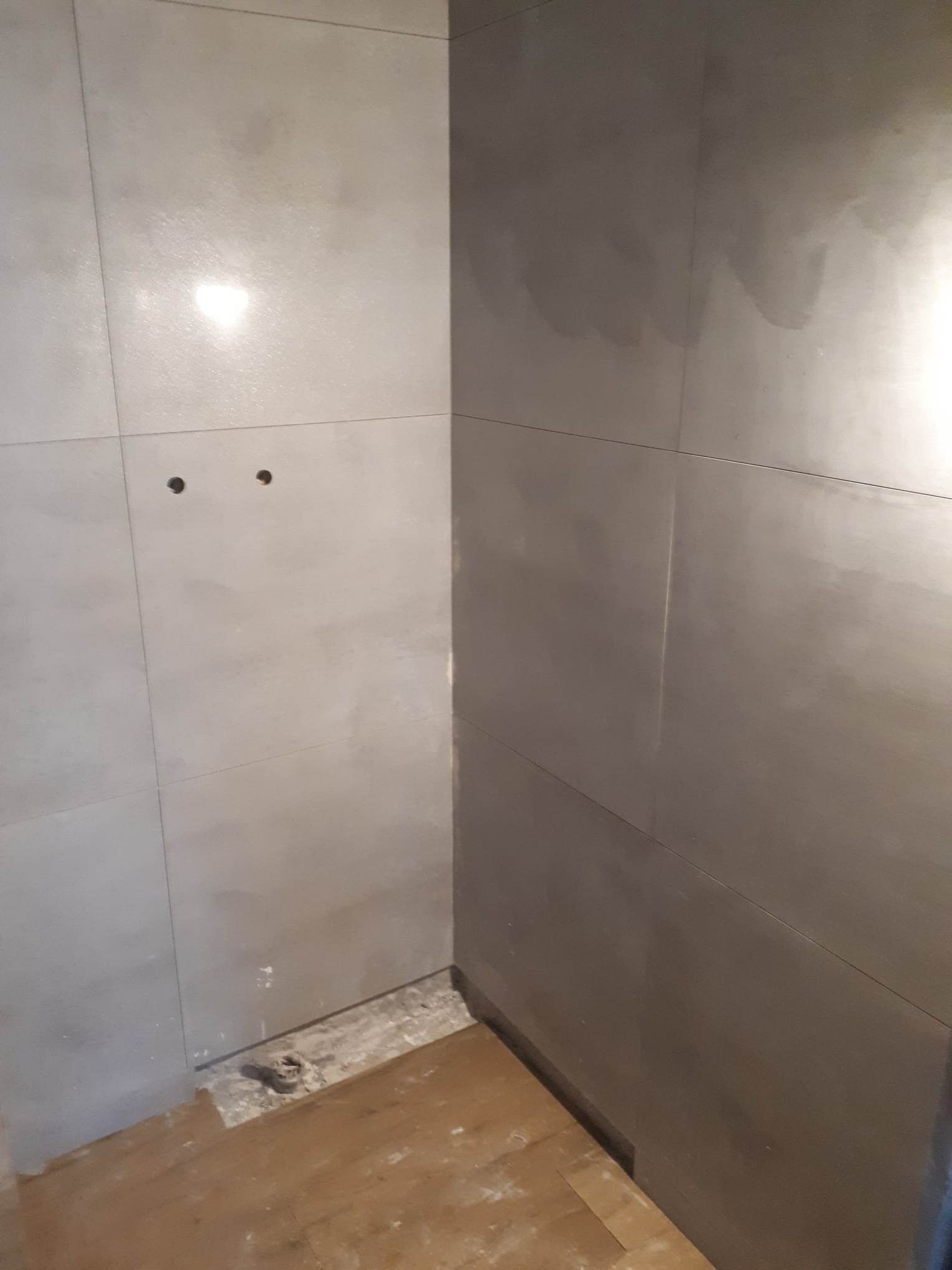 Fragment łazienki z szarymi, błyszczącymi płytkami ściennymi ułożonymi w poziomie, widoczne dwa otwory na armaturę i niedokończona podłoga z fragmentami płytek i kleju.