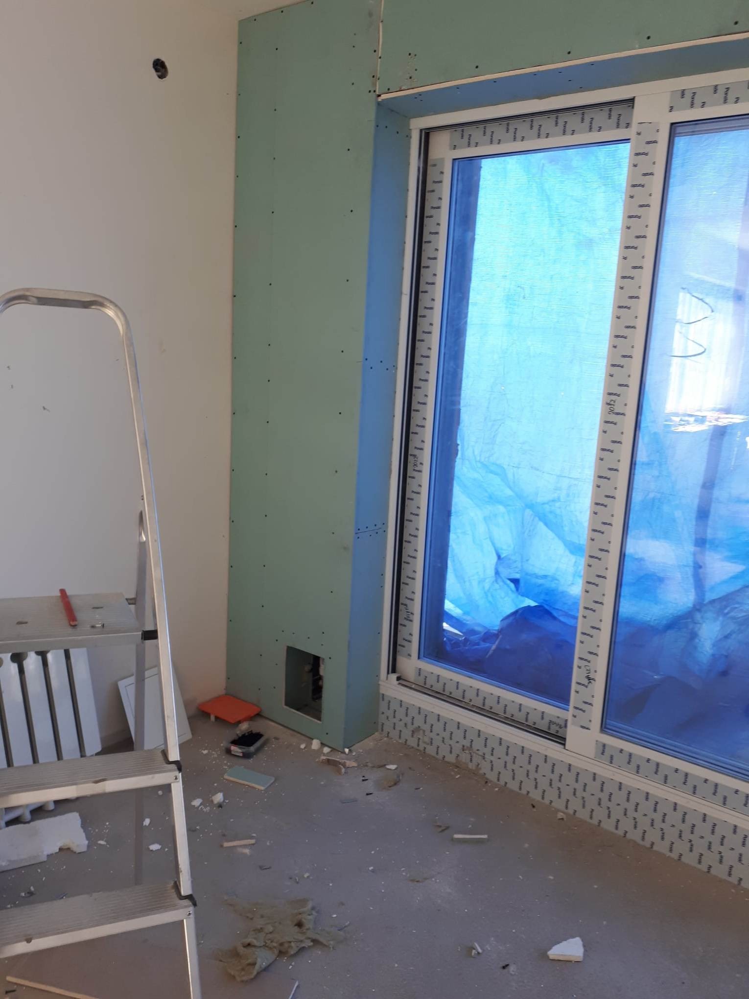 Wnętrze w trakcie remontu: okno zabezpieczone folią, ściana z płyty gipsowo-kartonowej, drabina aluminiowa i drobne odpady budowlane na podłodze.