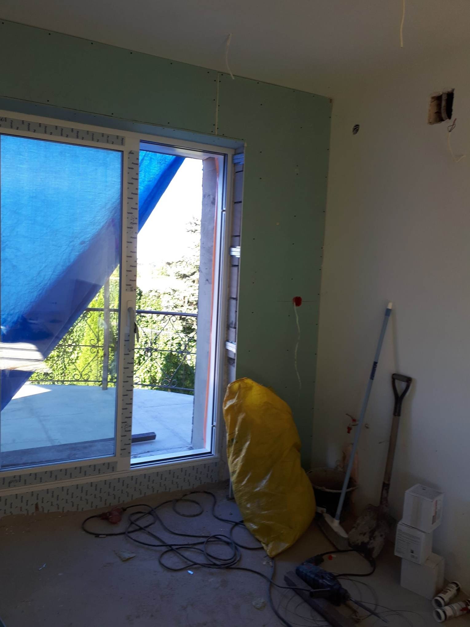 Wnętrze w trakcie remontu: ściana z płyt kartonowo-gipsowych, okno zabezpieczone folią, narzędzia i worki na podłodze.