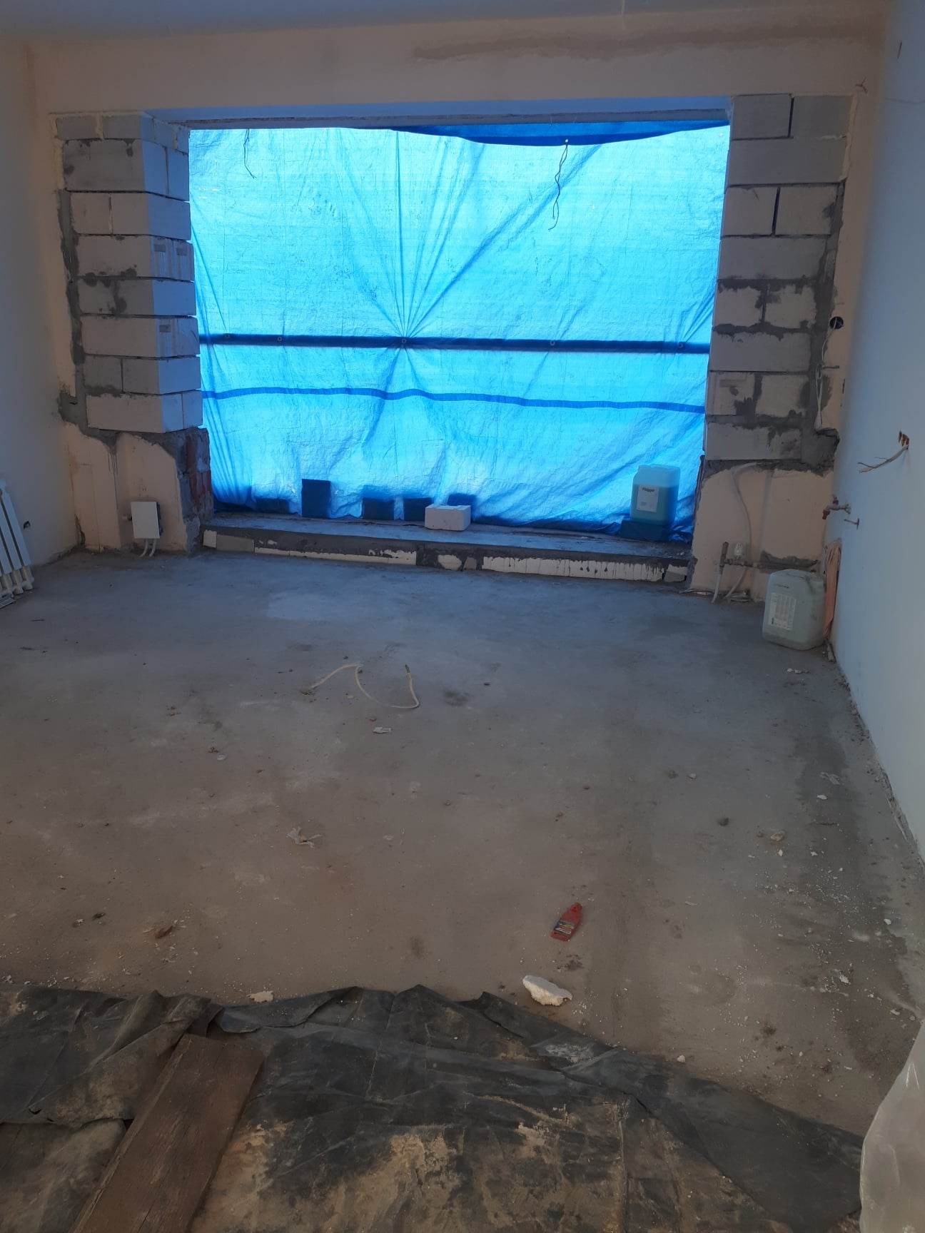 Wnętrze remontowanego pokoju z oknem zabezpieczonym niebieską plandeką, ściany z surowego betonu komórkowego, podłoga w trakcie wylewki, widoczne pojemniki z farbą lub gruntem i drobne narzędzia.