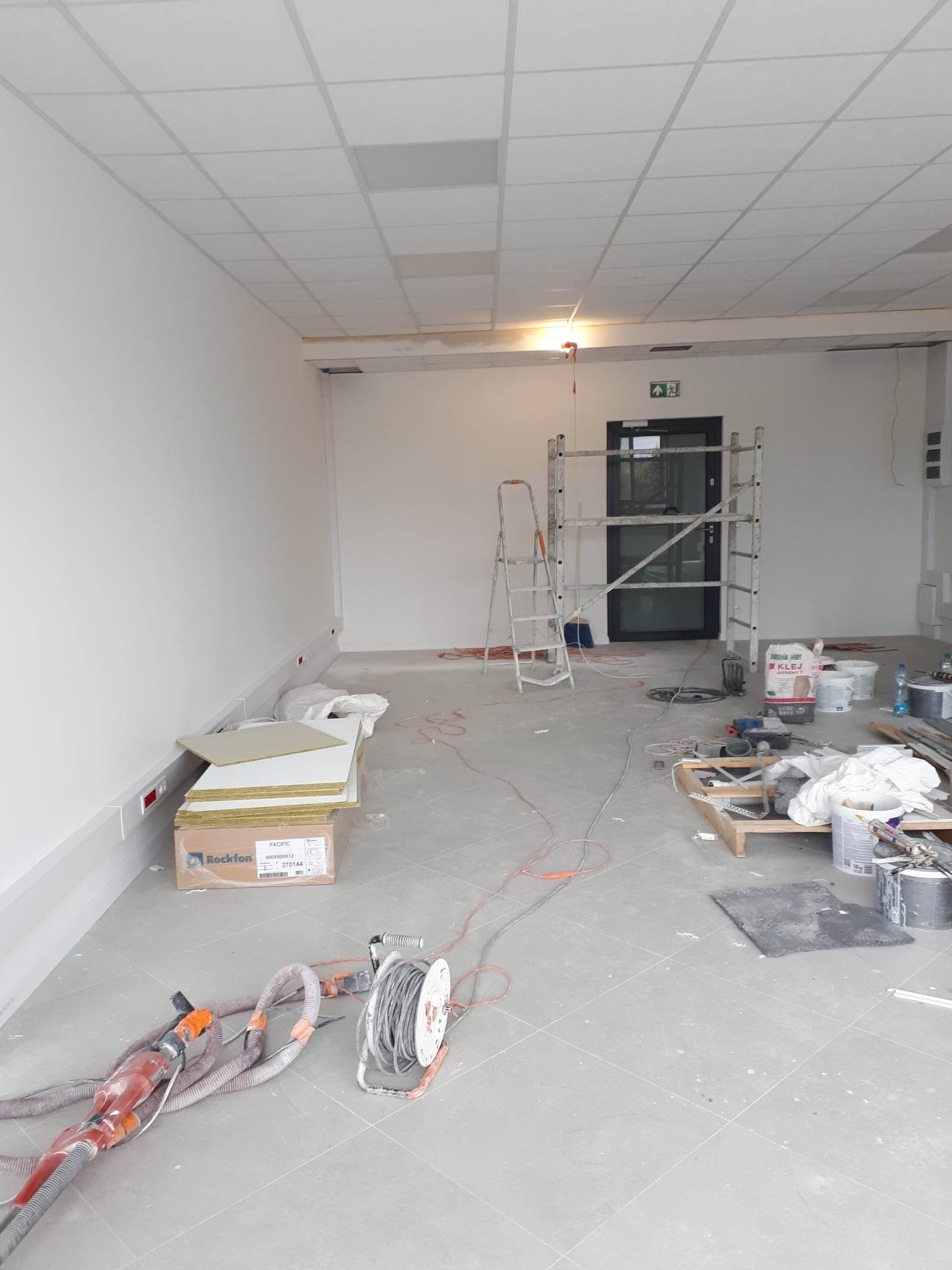 Wnętrze w trakcie prac wykończeniowych: białe ściany, sufit podwieszany, drabina, rusztowanie, materiały budowlane (wełna mineralna Rockfon), narzędzia i resztki farby na podłodze.
