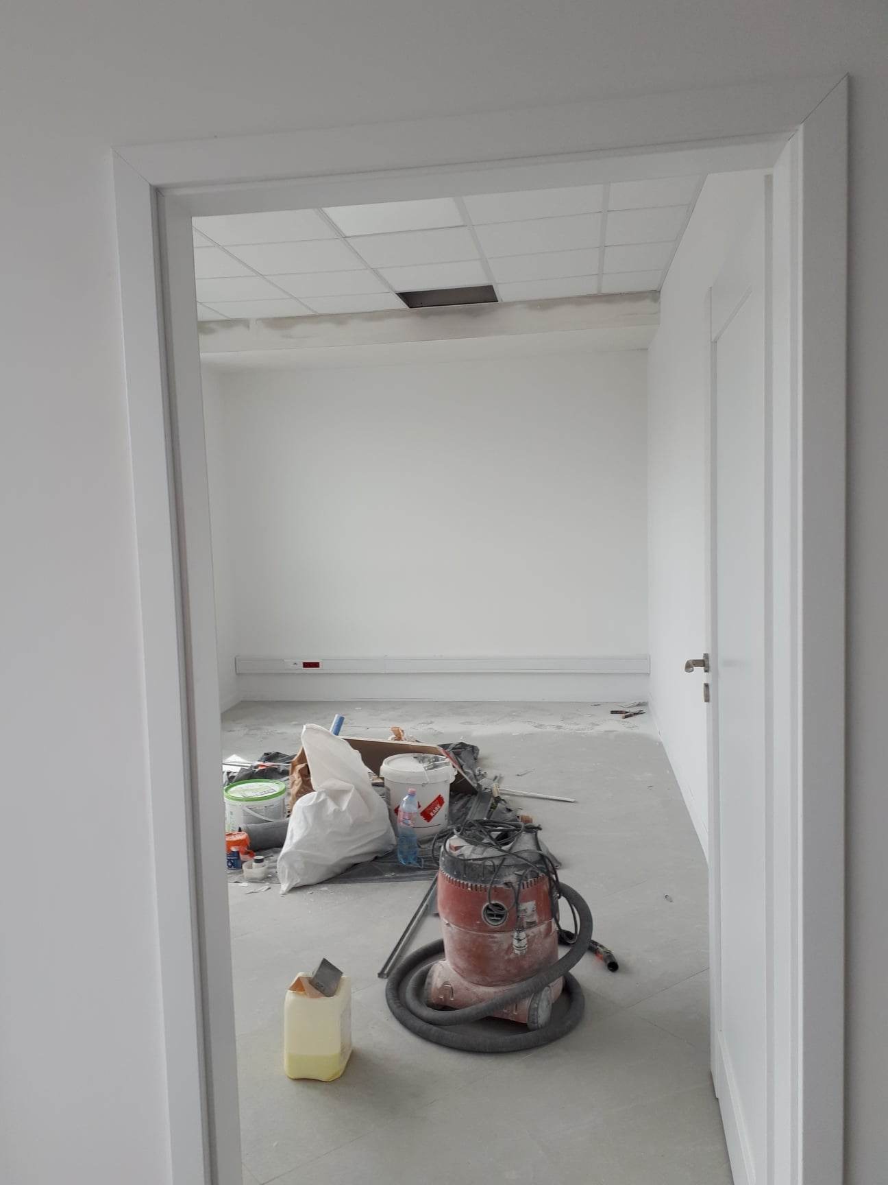 Wnętrze biura w trakcie malowania: białe ściany, sufit kasetonowy z otworem, widoczne narzędzia malarskie i odkurzacz przemysłowy.