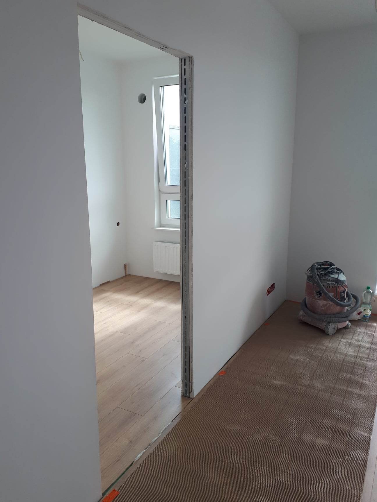 Wnętrze w trakcie remontu: świeżo pomalowane białe ściany, widoczny fragment podłogi z ogrzewaniem podłogowym i odkurzacz przemysłowy w rogu pomieszczenia.
