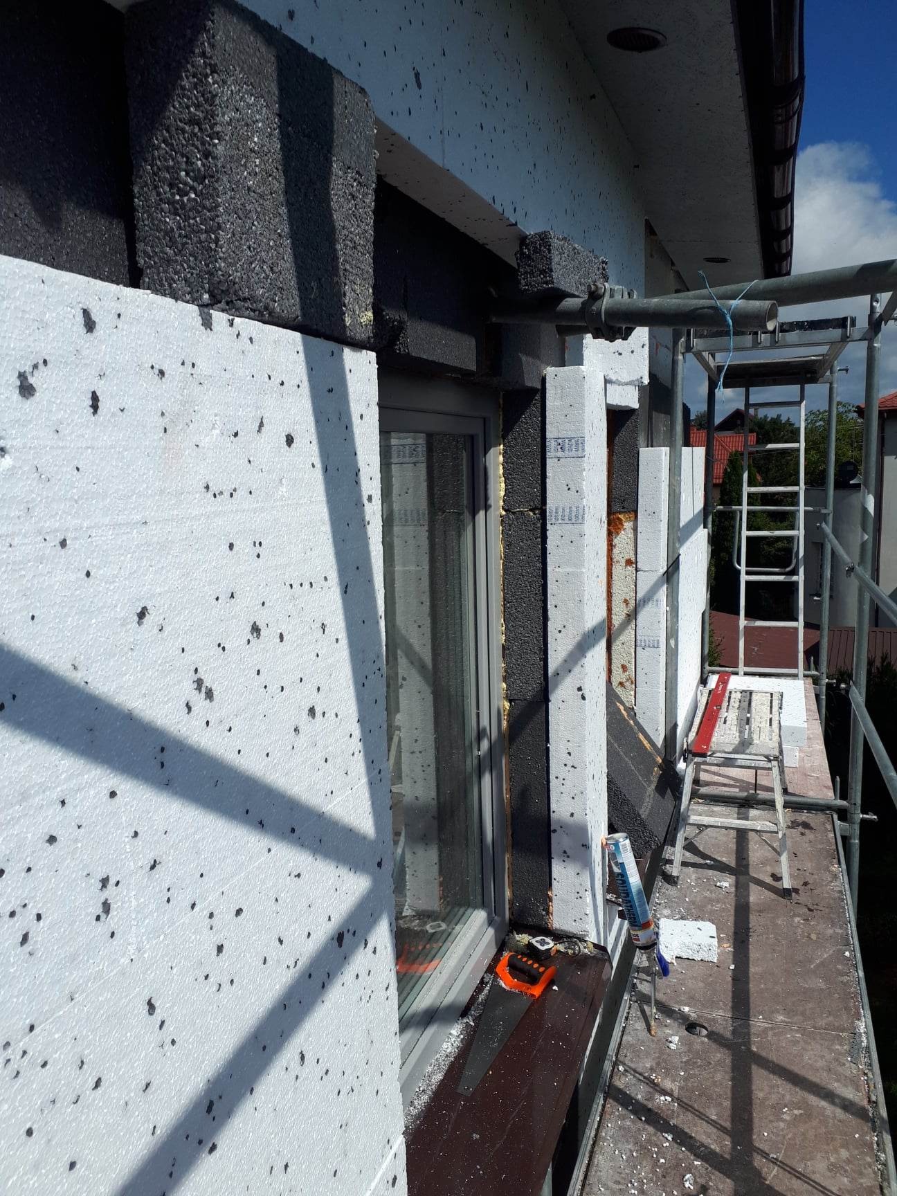 Ocieplanie elewacji budynku styropianem, widoczne rusztowanie, okno i narzędzia pracy.