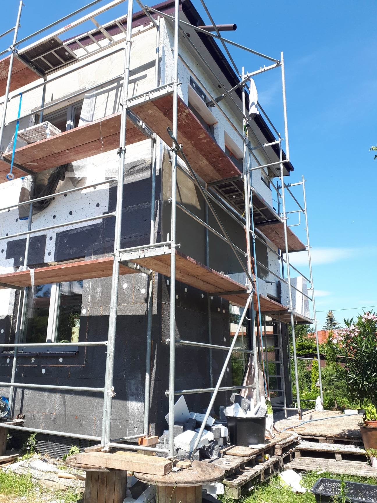 Dom w trakcie ocieplania, widoczne rusztowania, czarny styropian i szara warstwa kleju, elementy ocieplenia leżą na paletach przed budynkiem.